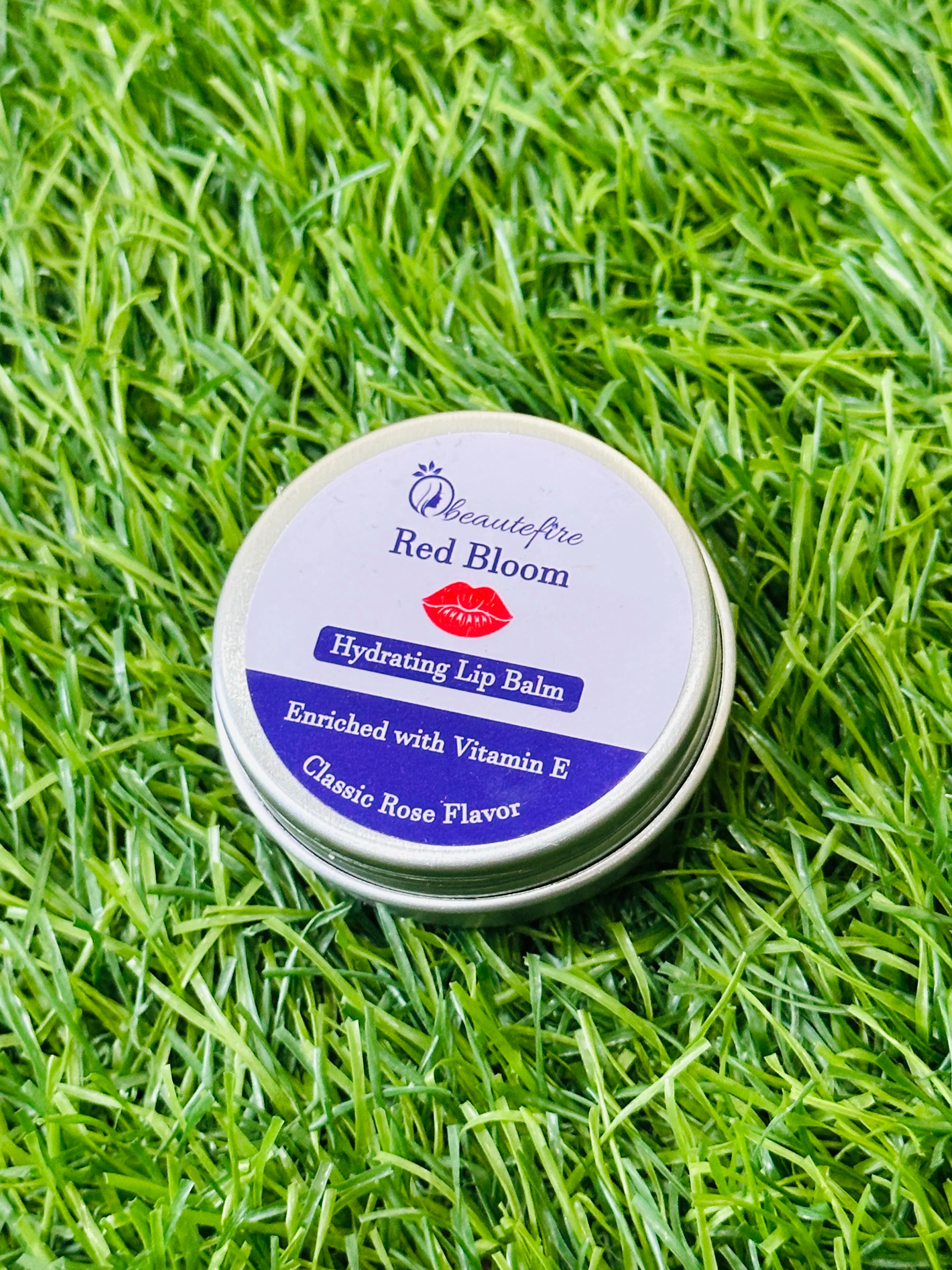 Beautefire Red Bloom Lip Balm placed on grass ā vibrant red nourishing balm captured in natural daylight.